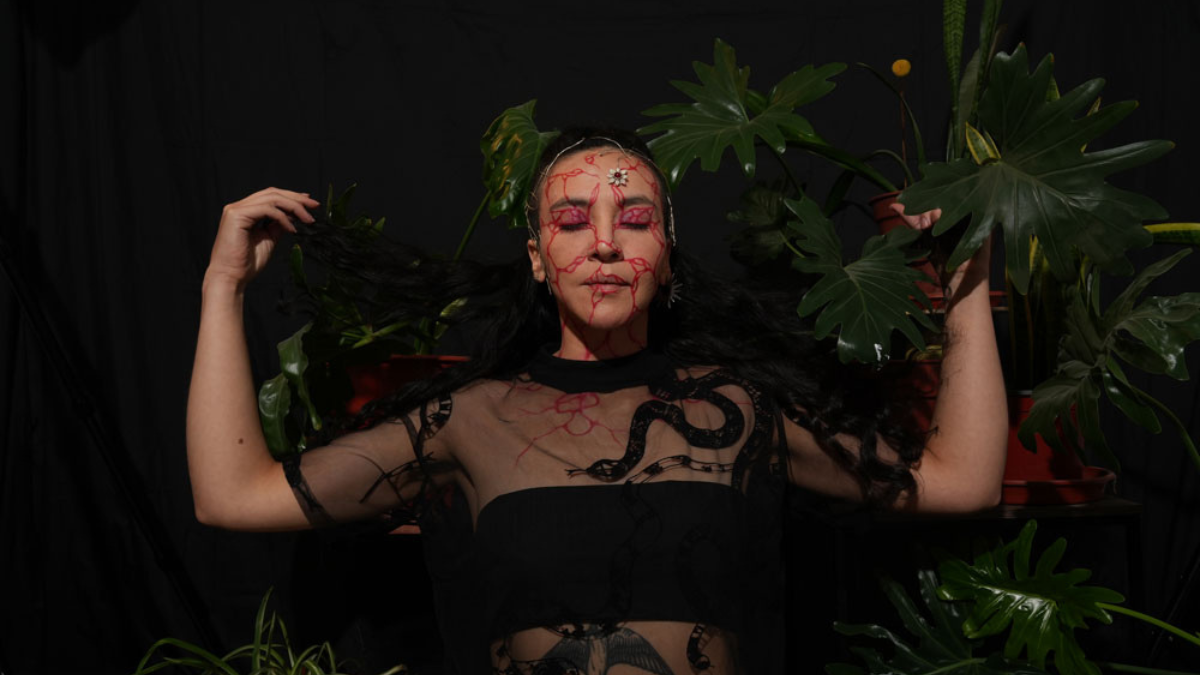 Cantautora chilena Paulina Pérez de TRAMA con maquillaje de raíces rojas y vestuario conceptual, rodeada de plantas en un fondo oscuro para Circulart 2026.