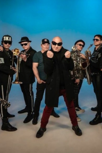 Fotografía de estudio de La Mosca Tse Tse, una banda de música ska compuesta por seis integrantes masculinos. El cantante principal posa en el centro con los puños cerrados, lentes oscuros y abrigo negro, flanqueado por músicos que sostienen instrumentos de viento como trombón y saxofón, sobre un fondo azul claro. Útil para ilustrar artículos sobre música ska, rock alternativo y cultura musical.